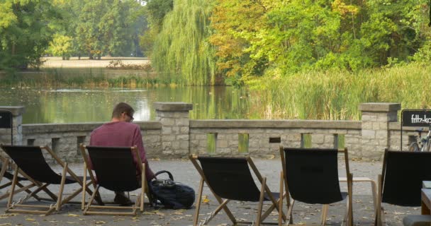 Hombre en camisa granate y pantalones beige se sienta relajado en la tumbona en la terraza frente al lago se pone de pie toma su mochila y se va — Vídeos de Stock