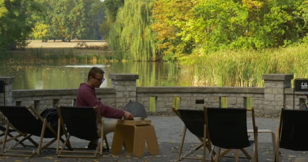 Człowiek bierze Tea Cup z tabeli w pobliżu jego krzesło i napoje z tarasu Cup naprzeciwko jeziora ptaki siedzi w dół na powierzchni wody Park Area — Wideo stockowe