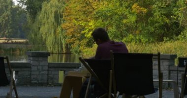 Adam Oturur ve Su Yüzeyine Bakar Çaydanlıktan Bardağına Çay Döker ve İçerir Sandalyesine Eğilir ve Arka Görüş Park Gölü'nü rahatlatır.