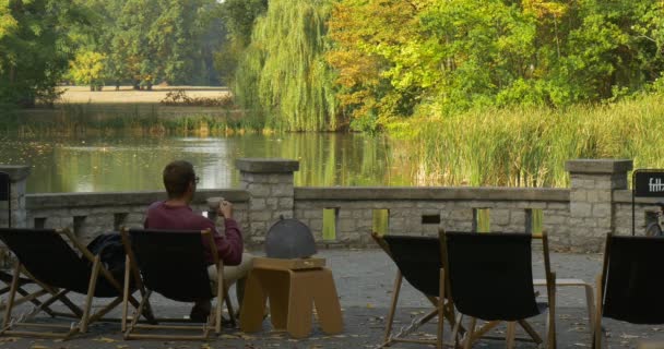 Mann sitzt entspannt im Liegestuhl er lehnt sich im Stuhl zurück und trinkt aus der Teetasse in der Hand Mann in kastanienbraunem Hemd und beiger Hose Parkgelände See — Stockvideo