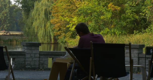 Muž sedí a dívá se na hladinu vody nalévá čaj z čajové konvice na pohár a nápoje je zpátky ve svém křesle a uvolní zpět pohled na jezero Park — Stock video