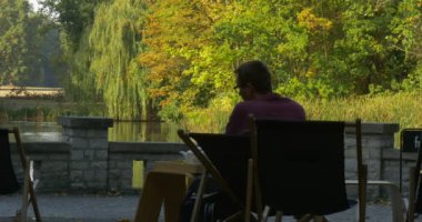 Elinde Fincan ile Man Oturur Sandalye de Arka Görünümü O Masaya Fincan koyar Onun Siyah Sırt Çantası Alır Ayağa Kalkar ve Uzak Park Alanı Göl Goes