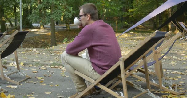 Az ember ül a Deckchair fehér kupa az ő jobb keze italokat a kupa majd úgy néz ki a távolság és meditates Man helyezi kupa félre, és úgy hátizsák — Stock videók