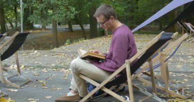Adam Şezlong oturur ve kitap okur sonra çay içecekler fincan alır ve Kamera Park Alanı Güneşli Sonbahar Günü bak
