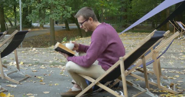 El hombre con el libro en la mano izquierda y la taza de té se sienta en la tumbona lee cuidadosamente el libro y bebe de la taza El hombre se inclina hacia atrás en su silla — Vídeo de stock
