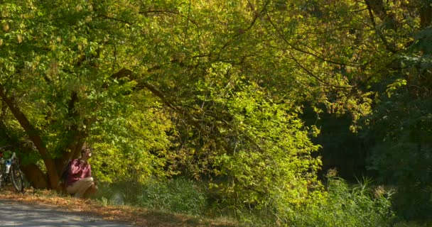 El hombre se sienta en la colina bajo el árbol verde que se extiende cerca del lago toma su mochila se levanta y mira el lago luego se aleja en su bicicleta — Vídeos de Stock