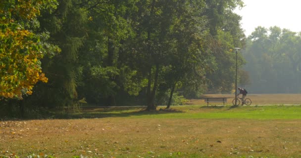 Uomo Stnads con bicicletta vicino alla panchina nel parco che ottiene sulla moto e viaggia lontano uomo in pantaloni beige e camicia marrone Sunny Autumn Day — Video Stock