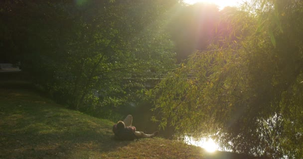 L'homme est allongé sur le sol à The Lake Bank Sun Rays Silhouette sac à dos est sous sa tête L'homme s'est levé secoue prend sac à dos et s'éloigne — Video