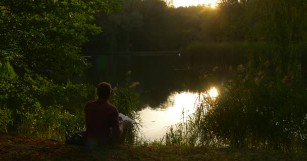 Homme avec ordinateur portable sur ses genoux s'assoit sur le sol au bord du lac Vue de dos, il regarde l'eau et imprime quelque chose sur le clavier L'homme vient à l'eau — Video