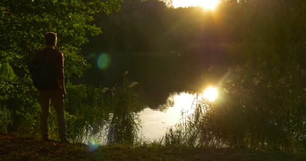 Homem chega ao lago se senta no chão e leva laptop de sua mochila ele abre LAptop e começa a trabalhar Lakeshore Sunset Autumn Day — Vídeo de Stock