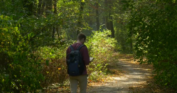 Man i rödbrun skjorta med ryggsäck på ryggen turist han går på vandringsleden i trä och läser boken på gå tillbaka se varma höstdagen — Stockvideo