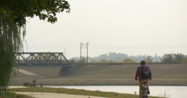 Homem com mochila está montando a bicicleta pelo beco do parque ao longo do rio sai da bicicleta de pé e olhando para o casal do rio está andando com o cão — Vídeo de Stock