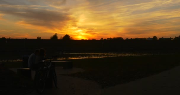 Koppel man en vrouw zitten aan de tafel silhouetten fiets is staand paar staat op wandelingen weg fel gele zonsondergang bij rivier kleurrijke wolken — Stockvideo