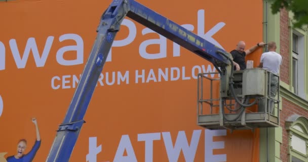 Dwóch mężczyzn pracowników w wieży Truck instalujesz pomarańczowy reklama sztandar w ramce na ścianie budynku wieży idzie w dół transparent z bliska — Wideo stockowe