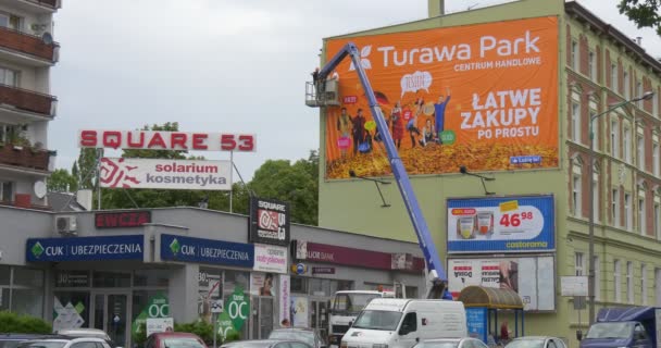 Работники грузовой башни устанавливают оранжевый рекламный плакат в металлическую рамку в красных кварталах здания городского пейзажа дорожных автомобилей ходьба людей — стоковое видео