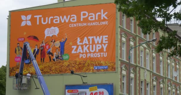 Deux hommes ouvriers dans la tour de camion installent une bannière publicitaire orange dans le cadre en métal aux blocs rouges construisant des fenêtres de bâtiment — Video