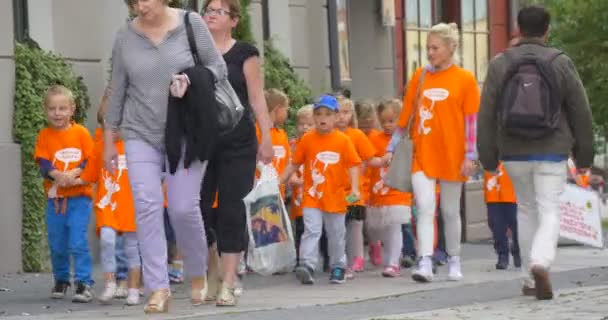 Les enfants en T-shirts orange marchent avec leur enseignant par les enseignants de la rue Les parents mènent les enfants vers la caméra Les voitures se déplacent de jour — Video