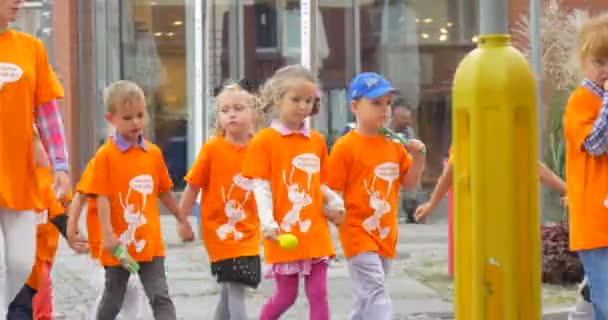 I bambini in arancione T-Shirt stanno camminando con il loro insegnante dagli insegnanti di strada I genitori stanno conducendo i bambini Le auto si muovono Negozi Finestre Di giorno — Video Stock