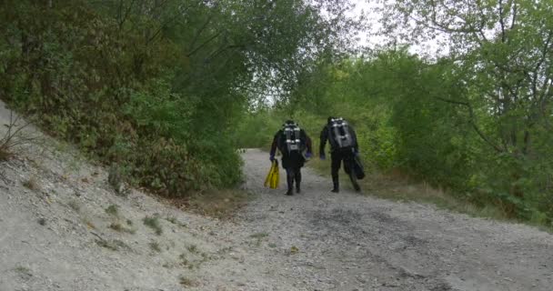 Dos hombres buceadores en trajes de baño están caminando por el sendero Árboles verdes a lo largo del sendero Los buceadores van a bucear Terminado el buceo al aire libre Otoño — Vídeos de Stock