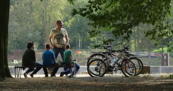 Les gens de la famille sont assis sur le banc dans le parc Les gens ont un repos Les vélos sont debout près des gens L'homme sur un vélo dans le casque est de passage — Video