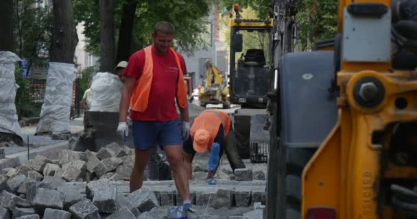 Werknemers in oranje werkkleding effenen de weg bij City Street weg reparatie gele graafmachine is permanent dicht bij Heap van blokken Transpors de blokken — Stockvideo