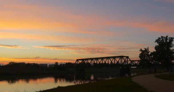 El hombre de blanco está sentado en la orilla La mujer camina a lo largo del estanque del río Ripeando las siluetas de los árboles de agua Reflexión Bright Sunset Pink and Yellow Sky — Vídeos de Stock