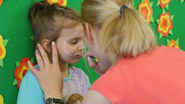 Educadora joven rubia está pintando la cara de una niña rubia pintando el hocico del gato en la cara de la niña por cepillo en el aula del jardín de infantes — Vídeo de stock