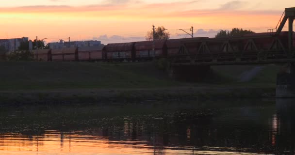 Güterzug Flussufer bewegt sich durch Brücke durch die Flussgebäude Stadt Dämmerung Sonnenuntergang Himmelsspiegelung im Wasser Wellen — Stockvideo