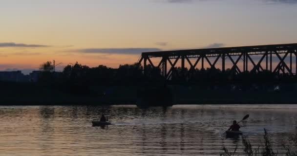 Dos hombres en los kayaks reman con paletas Personas Siluetas Río Banco Edificios Puente a través del río Puente Silueta Crepúsculo Puesta de sol — Vídeo de stock