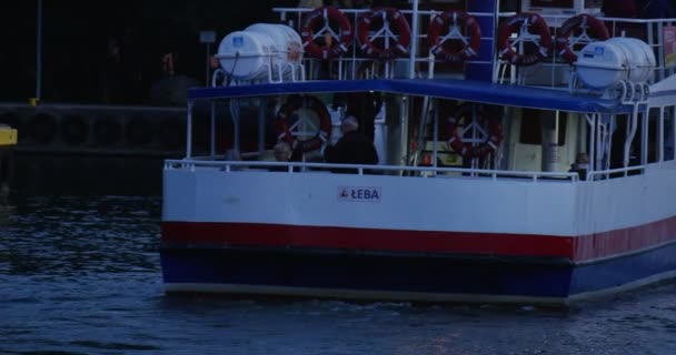 Barco a motor branco-azul-vermelho com pessoas na placa nada para o porto Fechar Visão traseira Pessoas caminham no cais Céu Nublado Noite de Verão Leba Polônia — Vídeo de Stock