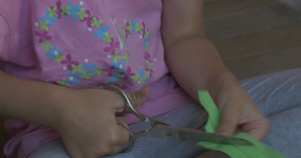 Petite fille en T-shirt rose avec tresse blonde est assis sur le sol, Faire une figure à partir de papier couleur, Couper les lignes du livre vert — Video