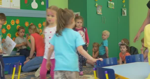 Группа счастливых детей с раскрашенными лицами бежит и стоит вокруг учителя в классе детского сада Дети идут танцевать — стоковое видео