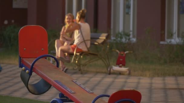 Mulheres, mães, estão sentadas no banco e conversando, Red Teeter-Totter em primeiro plano, Playground in the Yard — Vídeo de Stock