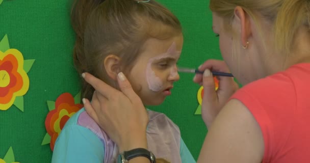 Pedagog młody blond kobieta jest malowanie twarzy trochę blond dziewczyna malarstwo pysk kota na twarz dziewczyny przez pędzel w klasie Przedszkole — Wideo stockowe