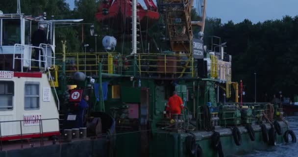 Зелёная строительная баржа с журавлем на борту плавает к портовому рабочему в оранжевом рабочем костюме на доске Люди идут по сумеркам на набережной — стоковое видео
