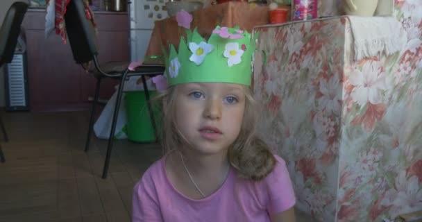 Niña en camiseta rosa con trenza rubia está sentada en el suelo, haciendo una figura de papel de color, lleva la corona de papel verde, hablando, gira — Vídeos de Stock