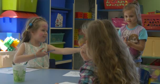 Dzieci dziewczynki i chłopiec siedzą przy stole i malarstwo uśmiechający się mówić Grupa szczęśliwy dzieci z pomalowanymi twarzami grają w klasie — Wideo stockowe