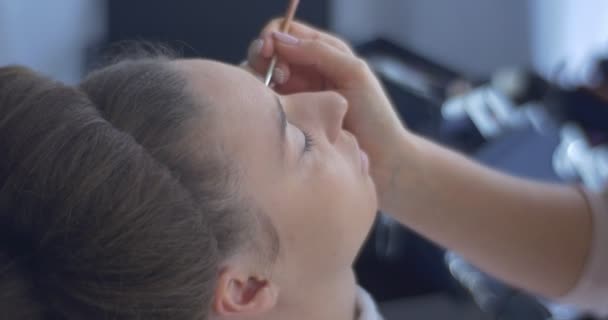 Stylist schoonheidsspecialiste is het maken van een oog make-up vrouw met kapsel krullen vrouw met bruine haren zit op een stoel schoonheidsspecialiste handen — Stockvideo