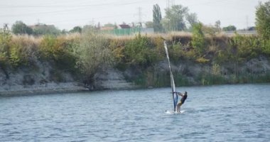 Yelken ile Windsurf Kurulu Man Sandy Bank Man boyunca Sulu Yüzey tarafından Hızlı Yüzen Windsurf Kostüm River Yeşil Ağaçlar Küçük Dalgalar Gökyüzü