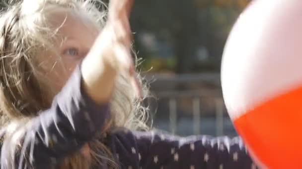 Petite fille aux cheveux longs et blonds lève les mains Jouer à la balle lance la balle attrape la balle La fille sourit Saut Parc Arbres Clôture Journée ensoleillée — Video