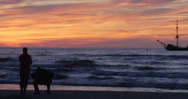 Människor familjer silhuetter män med ryggsäckar tar foto människor går genom sandstrand simning människor ljusa solnedgången vågor på havet — Stockvideo