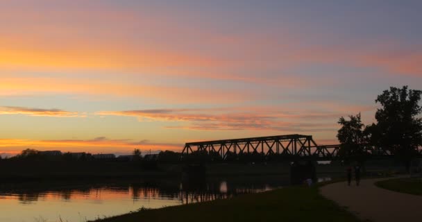 El hombre de blanco está sentado El hombre joven y la pareja de mujeres caminan El tren se mueve por el puente Siluetas de los árboles del río Bright Sunset Cielo rosa y amarillo — Vídeos de Stock