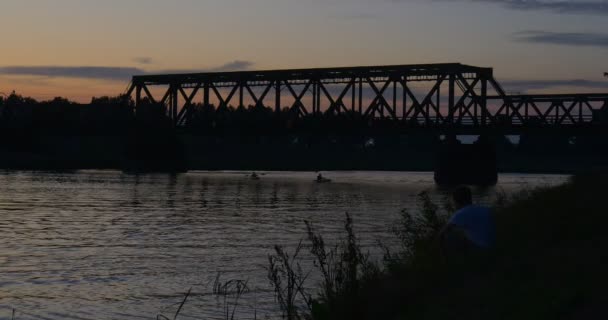 El hombre de blanco está sentado en la orilla del río El hombre deja a los hombres en el estanque del río Kayaks Ripeando las siluetas de los árboles de agua Reflexión Sunset Pink Sky Bridge — Vídeos de Stock