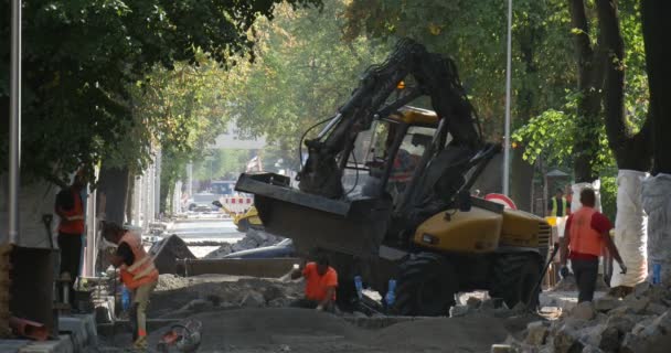 Los hombres trabajadores en ropa de trabajo amarilla en la calle Sity están cavando la excavadora amarilla Reparación de carreteras Edificios Casas Árboles verdes en segundo plano Señal de tráfico — Vídeo de stock