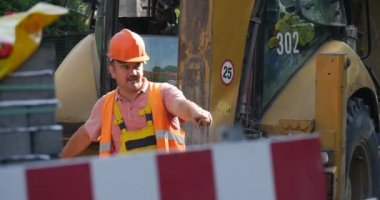 Workers in Yellow Workwear And Helmet Yellow Excavator Driver is Moving the Load by Scoop Road Repair Paving Road Safety Red and White Fence Sunny Day