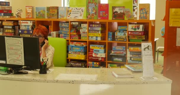 Mulher se senta no computador e fala sobre o celular bibliotecário menina com livro vem para a mesa João Paulo II Biblioteca Pública Municipal em Opole Polônia — Vídeo de Stock