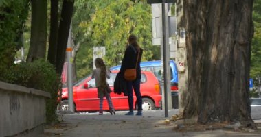 Kick Scooter Anne ve Kızı Anne ve Küçük Blonde Kız Street Alley İnsanlar Durur ve Yine Tree Trunks Arabalar Yürüyüş vardır