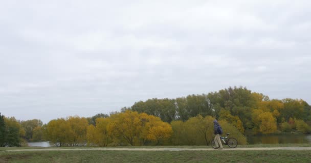 Uomo in Caldo Cappello Ushanka Cappello con Paraorecchie in pelliccia Uomo in Caldo Giacca Uomo con Zaino sta conducendo la pista ciclabile al Lago Autunno Giallo Alberi Cielo — Video Stock