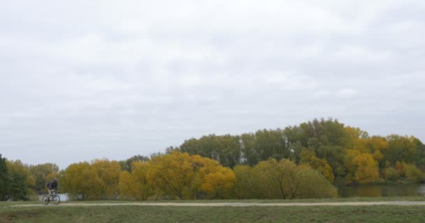 Hombre en sombrero caliente, sombrero de Ushanka con solapas de piel de oreja ciclista Hombre en chaqueta caliente Hombre con mochila está montando bicicleta sendero en el lago árboles amarillos otoño — Vídeos de Stock