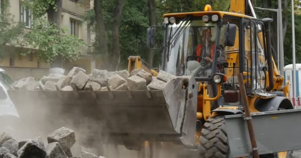 Föraren i gula grävmaskin griper block med Scoop transporter blockerar vägen reparation City Street arbetarna i Orange arbetskläder människor gå från Street — Stockvideo